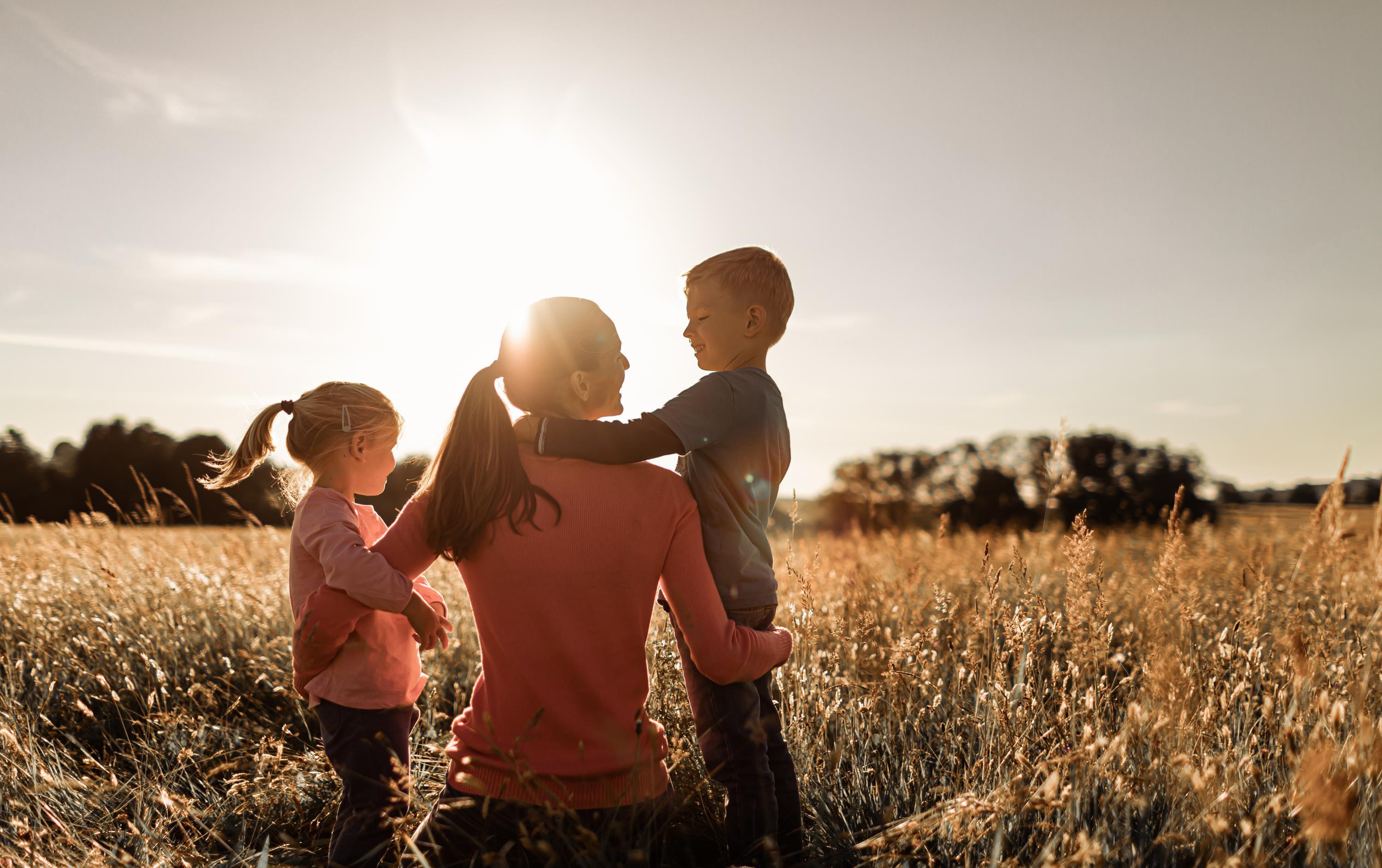 Mutter mit Kindern Mutter sitzt mit Tochter und Sohn im Arm in Wiese im Sonnenschein