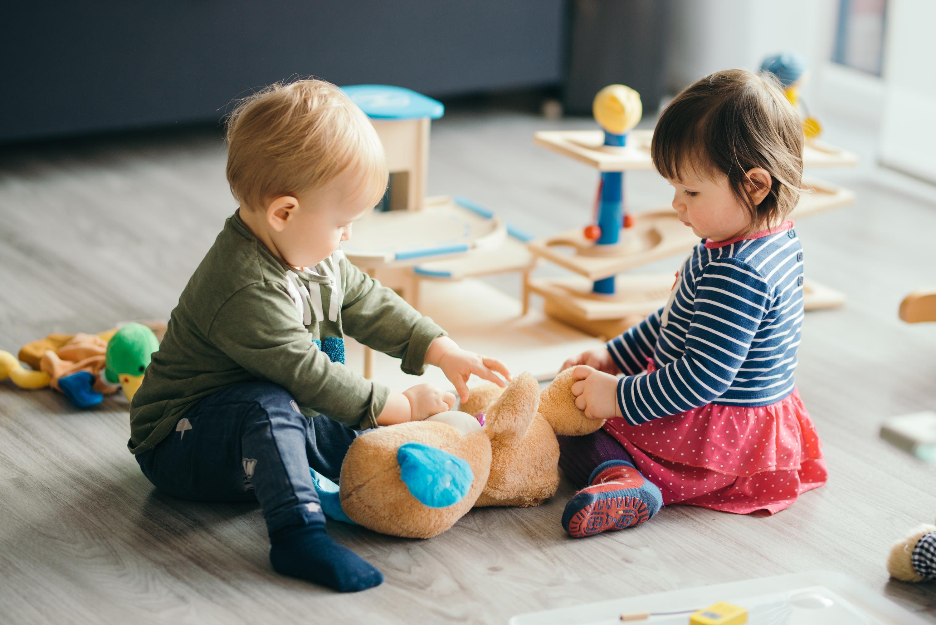 Spielende Kinder 2 Kleinkinder sitzen am Boden, spielen gemeinsam mit einem Stofftier
